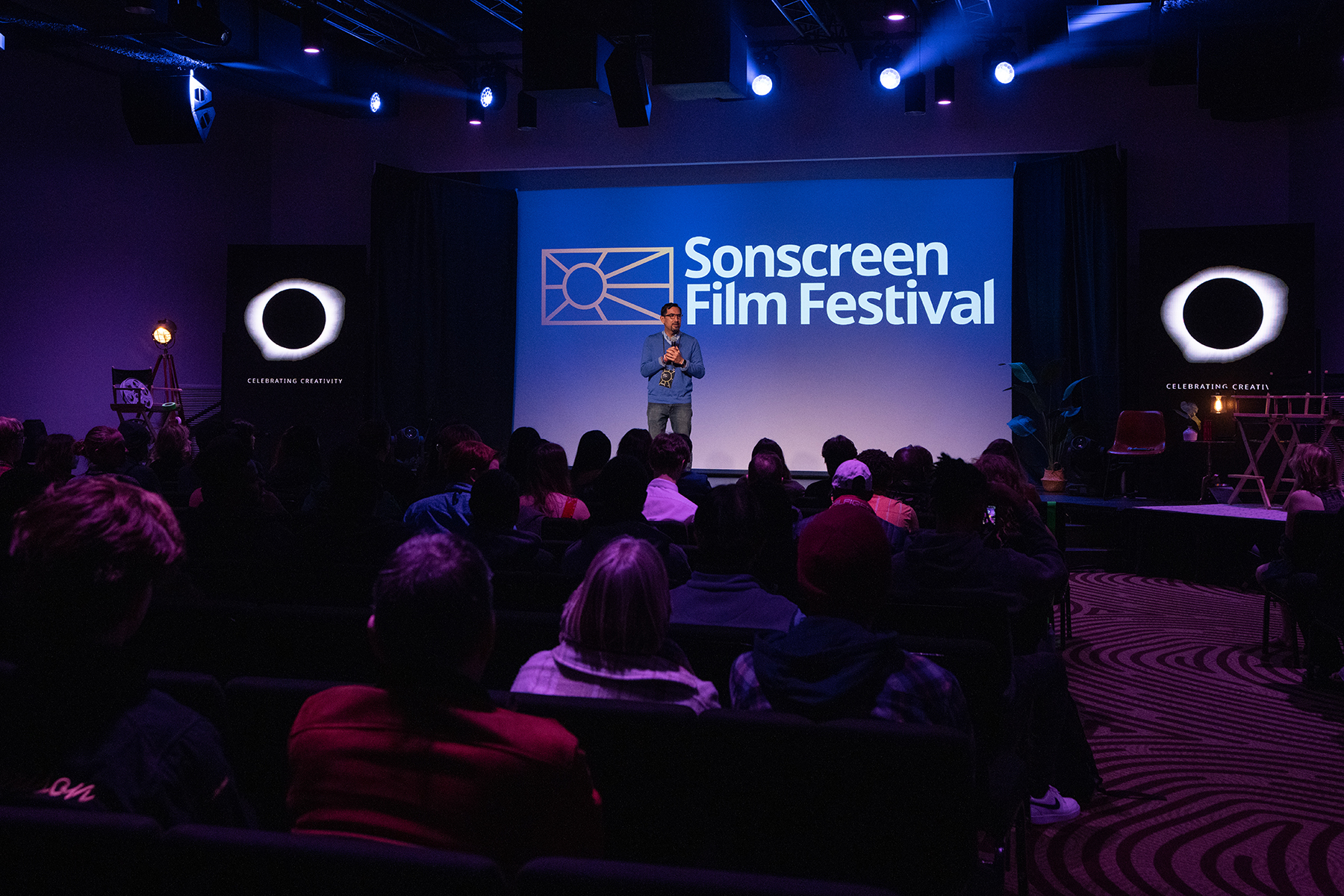 A Hispanic man stands in front of a screen reading "Sonscreen Film Festival." An audience can faintly be seen in a darkened room.