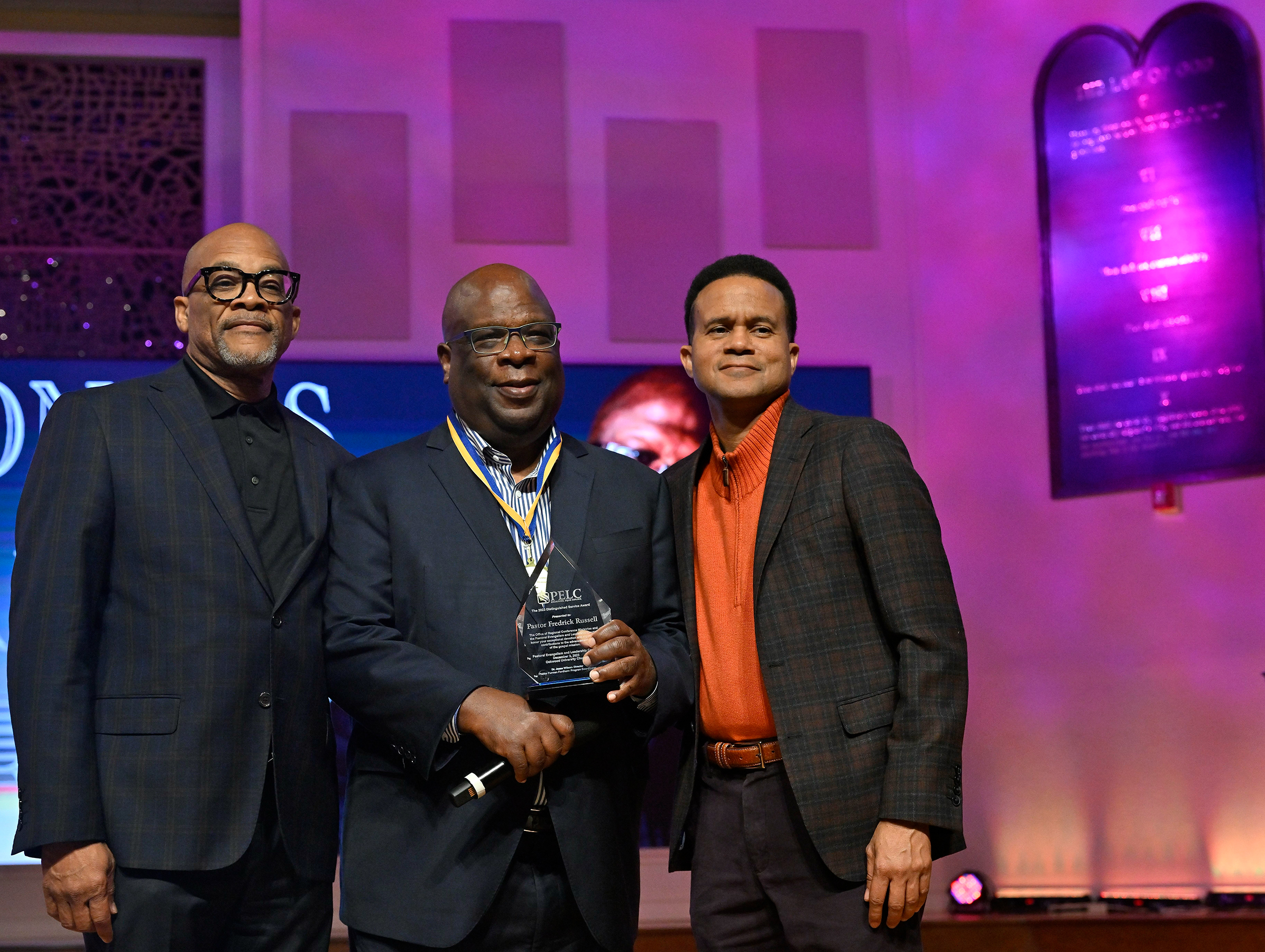 Three Black men on stage in a church. The one in the middle is holding an award.