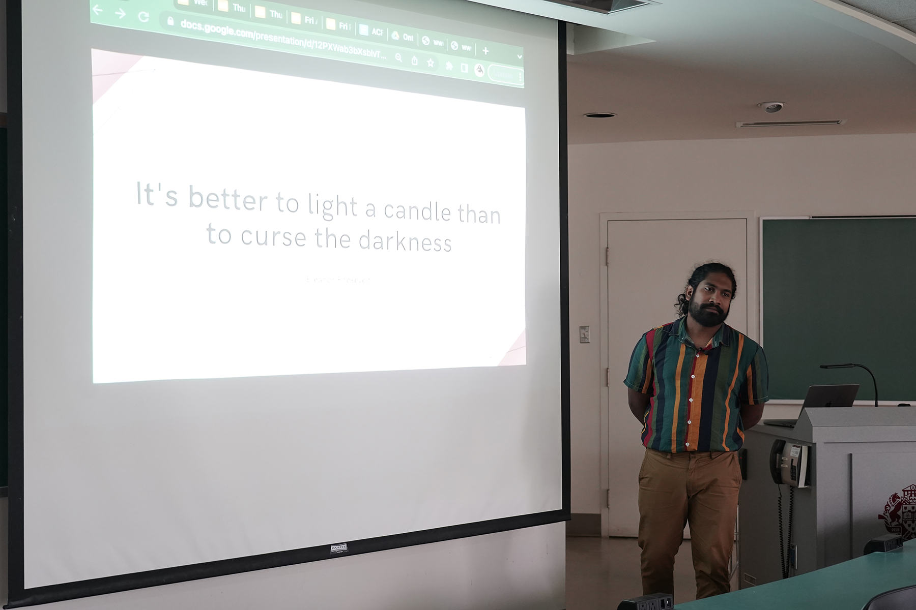 Brown man beside a screen, giving a presentation in a classroom.