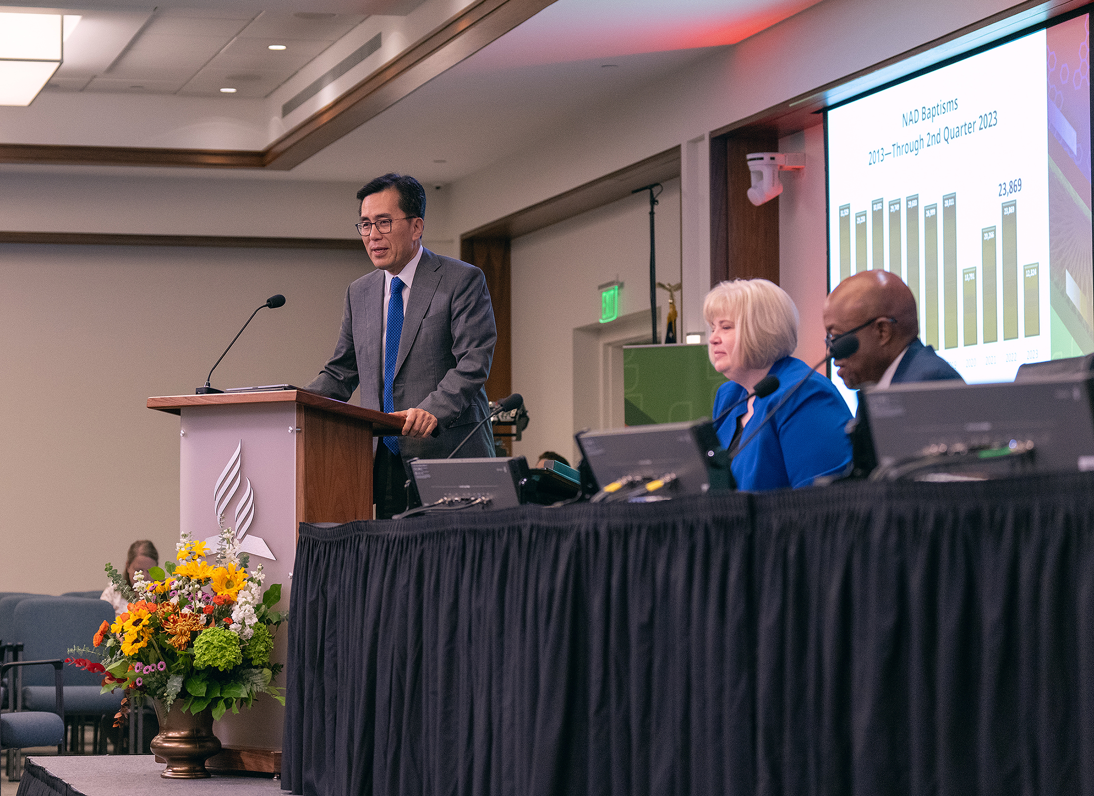 Asian man standing and speaking at a podium as white woman and black man look on.