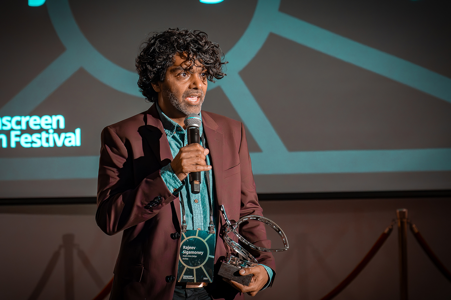 A brown man with curly hair holds an award that looks like a film reel and speaks into a microphone, emotional.