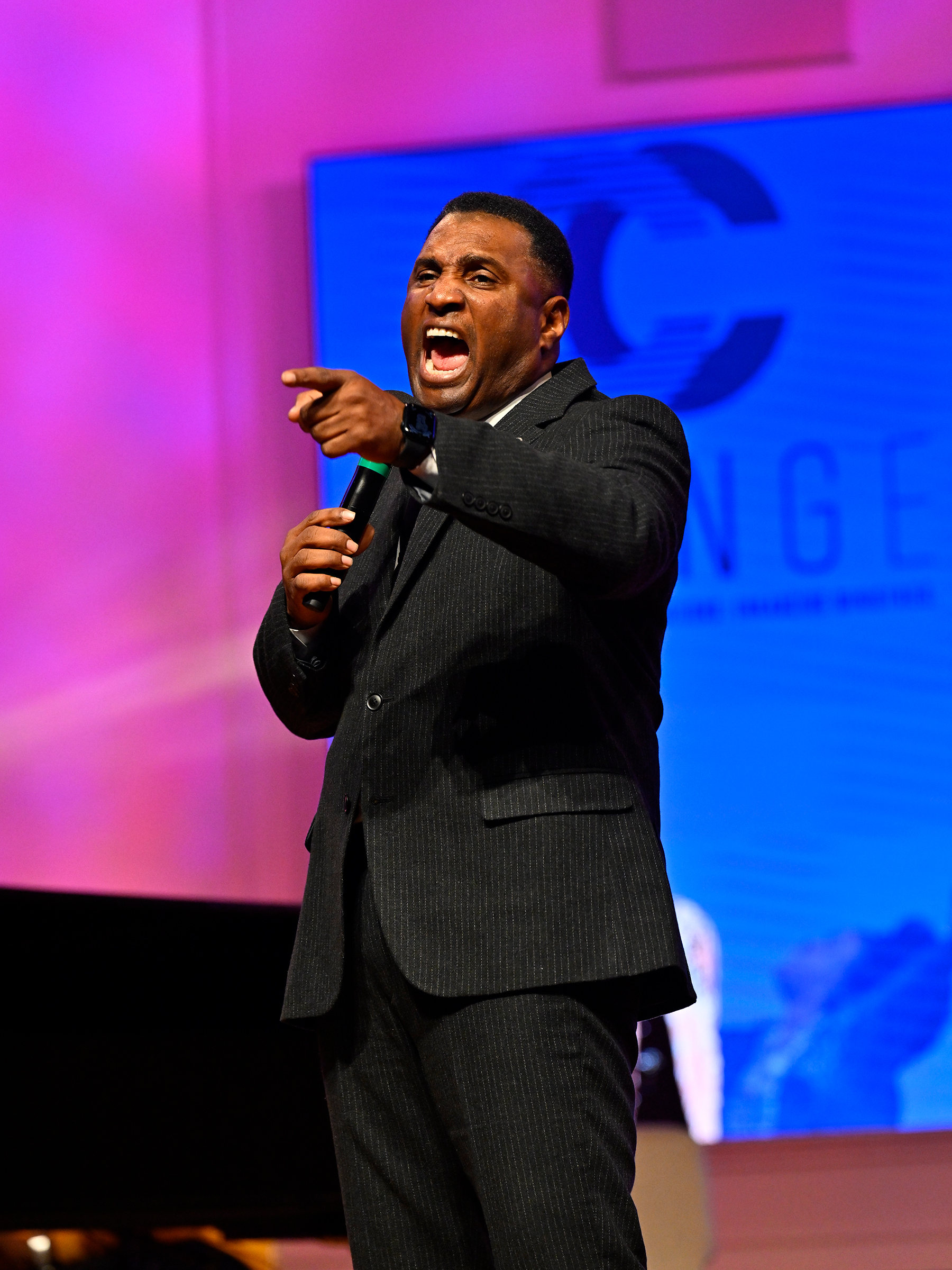 Black man on stage preaching, with his arm out and finger pointing.