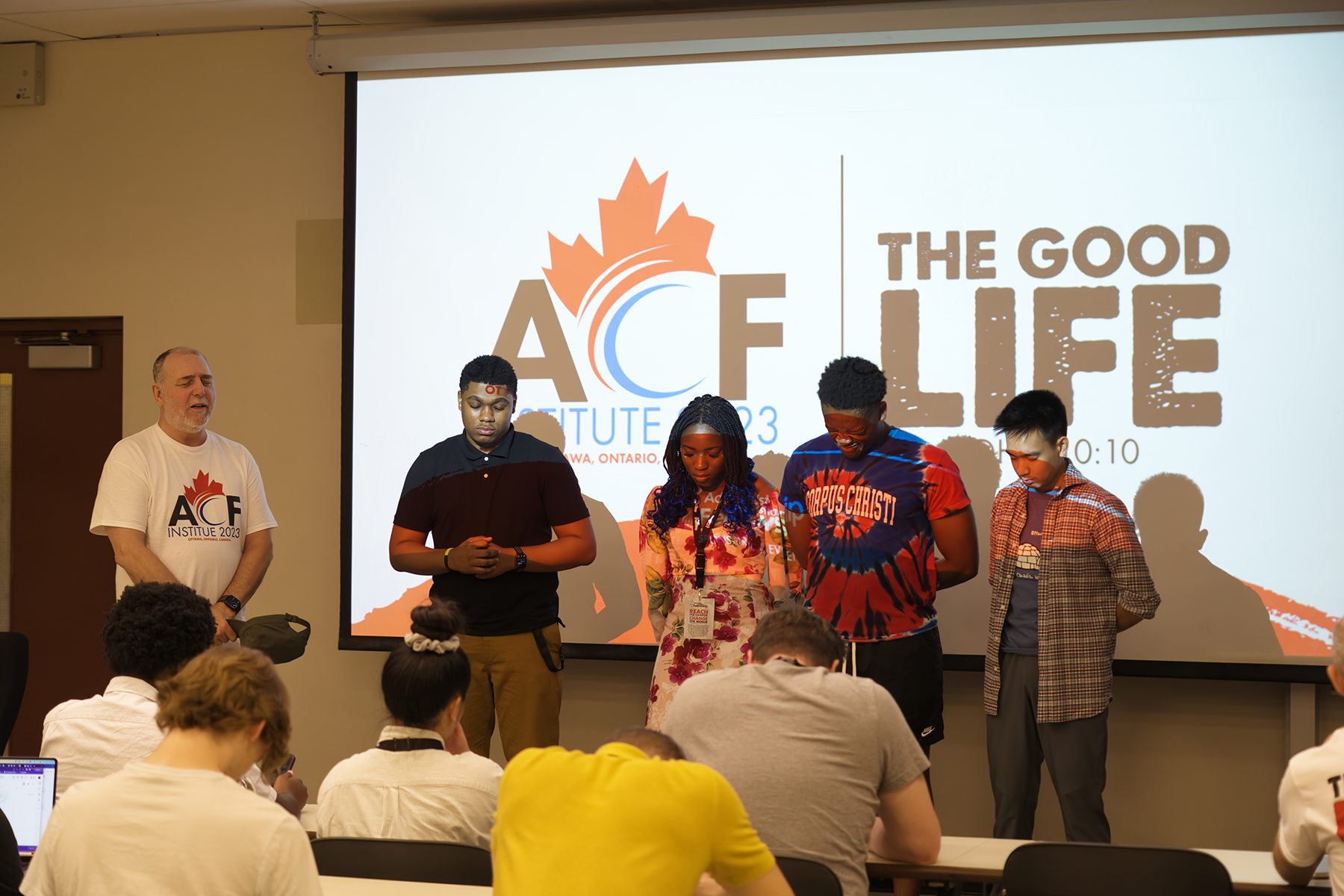 A diverse group of people praying in front of a screen reading "The Good Life."