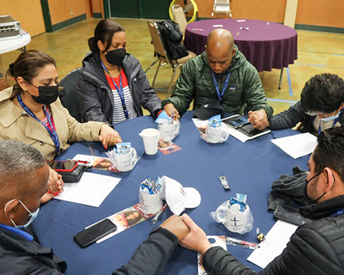 Six people praying, holding hands while seated at a round table.