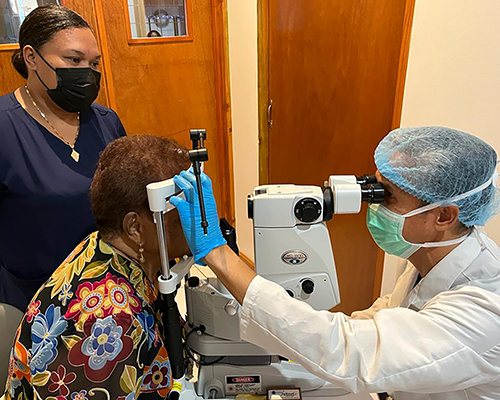 A woman in nursing uniform stands by while a man in doctor's coat gives another woman an eye exam