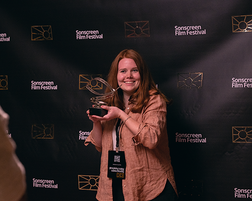 A young woman holding an award and standing in front of a media wall