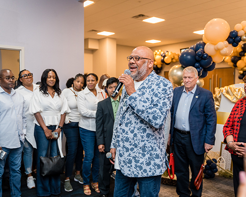 A photo of a man speaking at a celebratory event, with a crowd of people behind him.