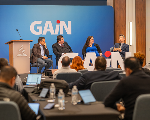 Left to right: two white men, a white woman, and an Asian man seated, are participating in a panel discussion as a conference. Onlookers are visible in the foreground