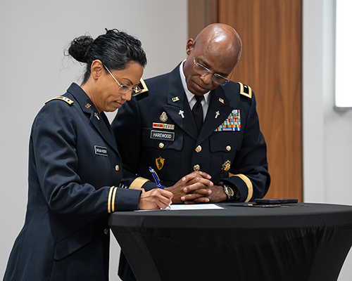 Lady with tanned skin signs a document as a black man watches; both are in military uniform