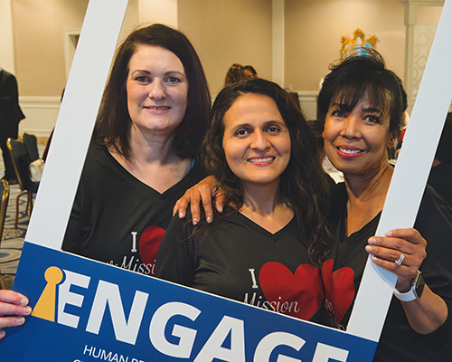 three women of different ethnicities in a cardboard frame reading "Engage"