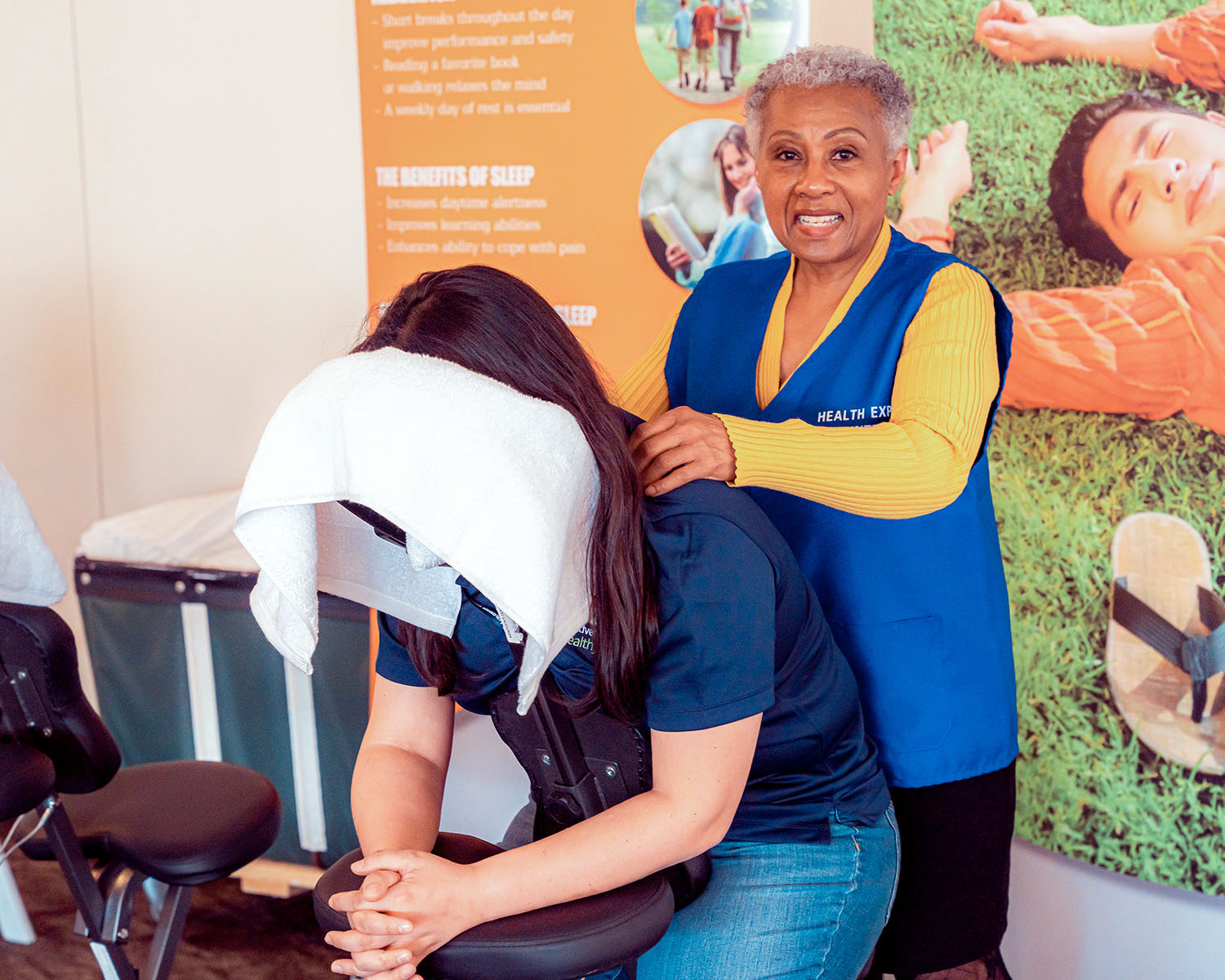 Photo of a smiling woman massaging the shoulders of another woman in a professional chair. The client's head is covered with a towel