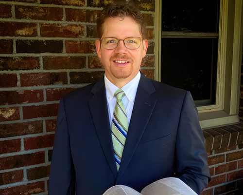 White man standing in front of a brick wall with a window, holding a Bible