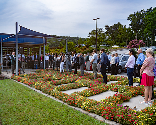 A man speaks under a tent to a small crowd outside along a path; there are video cameras pointed in his direction