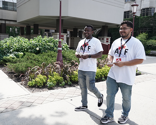 Two young men, one black (left), one Hispanic (right) walking down the street smiling with flyers in their hands. Both are wearing jeans and white t-shirts reading "The Good Life."