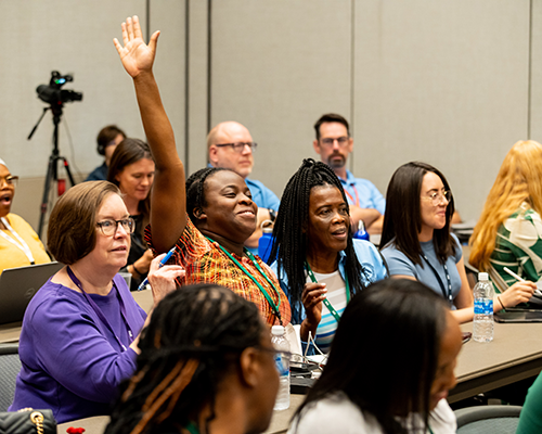 A black woman in a classroom smiles and raises her hand high. She's surrounded by other people, male and female, of diverse backgrounds.