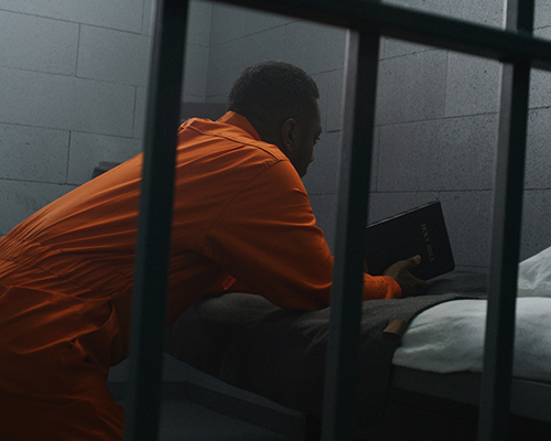 stock photo of man in prison reading Bible while kneeling in cell