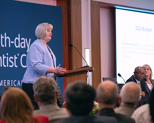 Smiling white woman standing behind a podium speaking to a crowd.