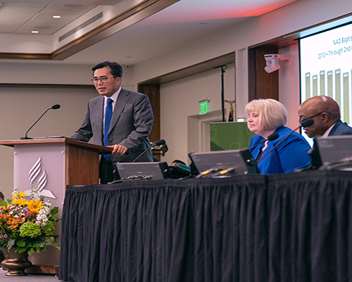 Asian man speaking at a podium, as white woman and black man (seated) look on. 
