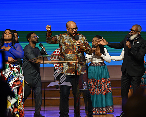 A Black praise and worship team on stage, wearing African outfits and singing passionately. The leader is in the middle.