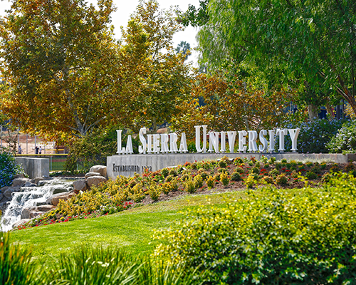 a photo of a sign reading "La Sierra University" surrounded by trees, green grass, plants, and a waterfall. 