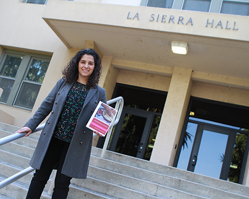 Hispanic woman standing in front of a building with the name "La Sierra Hall" holding a book. 