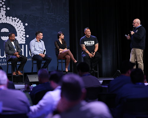 Panel of people sitting on a stage, left to right, black man, white man, white woman, and Samoan man, sitting. Their interviewer, standing, is a white man. 