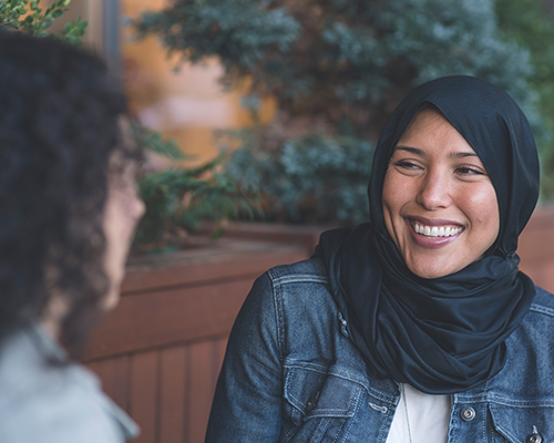 Muslim woman in scarf smiling at another woman whose face is obscured.