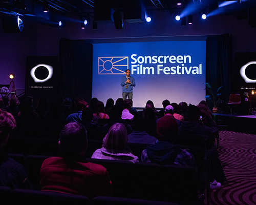 A Hispanic man stands in front of a screen reading "Sonscreen Film Festival." An audience can faintly be seen in a darkened room. 