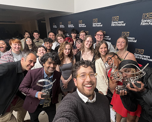 A diverse group of people stand in front of a media wall, some holding awards that look like a film strip