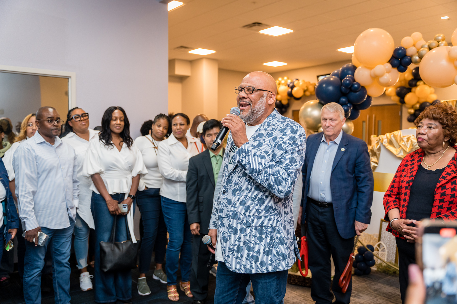 A photo of a man speaking at a celebratory event, with a crowd of people behind him