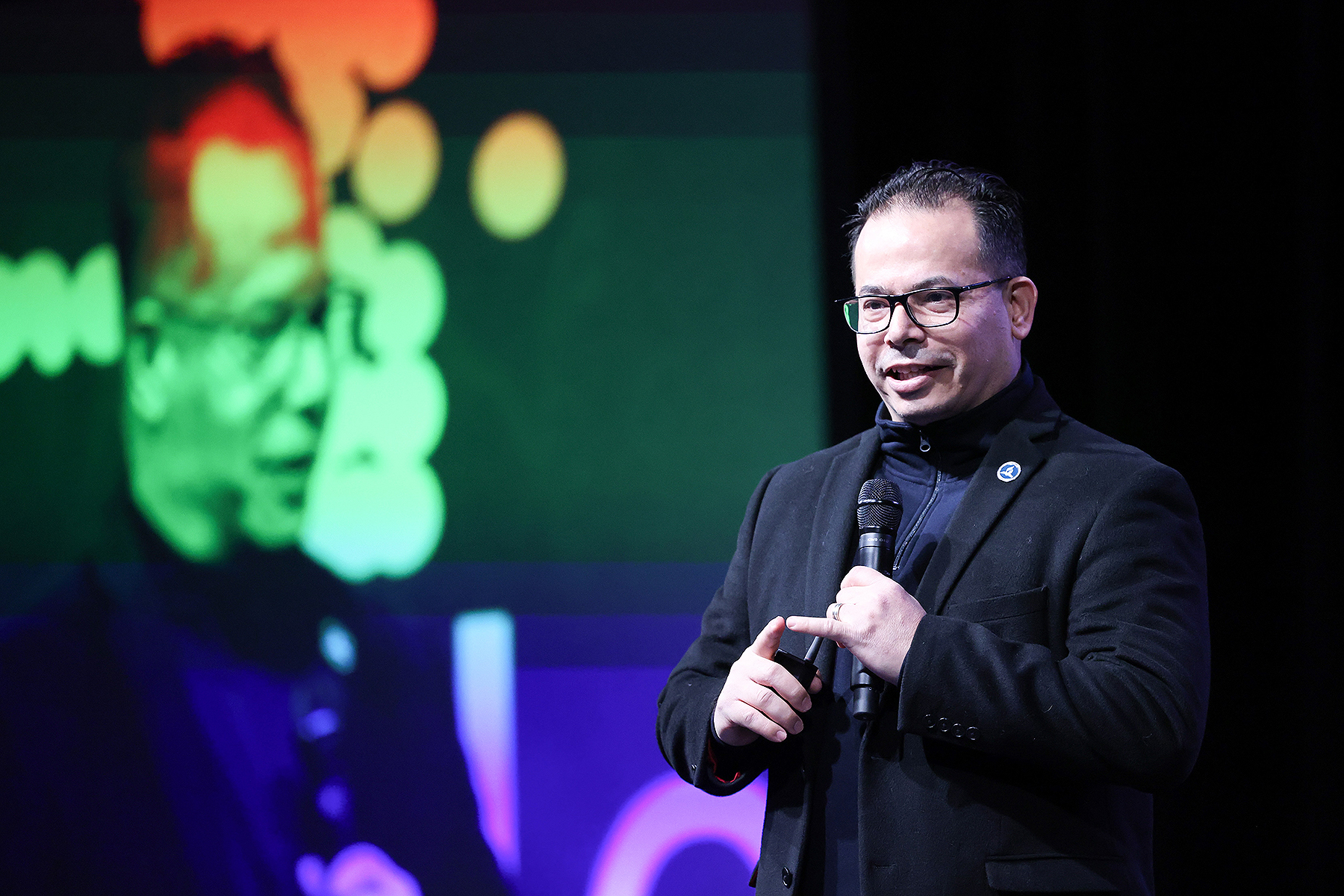 Hispanic man giving a presentation on a stage. Behind him is a blurry, colorful image of him speaking. 