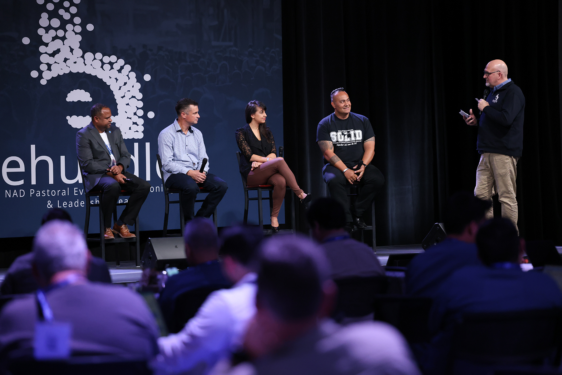 Seated on a stage, black man, white man, white woman, and Samoan man. Interviewer, standing, is a white man. 