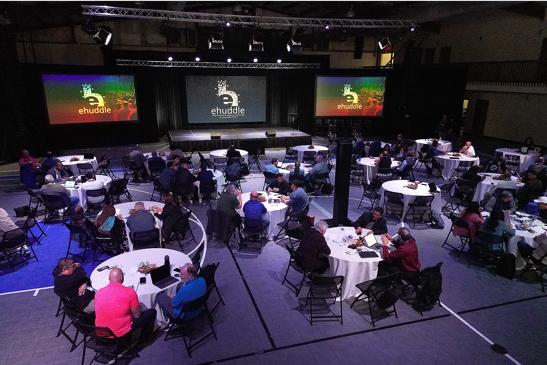 Wide-angle photo of a gym-turned-conference room, with screens at the front reading eHuddle and people seated at roundtables talking. 