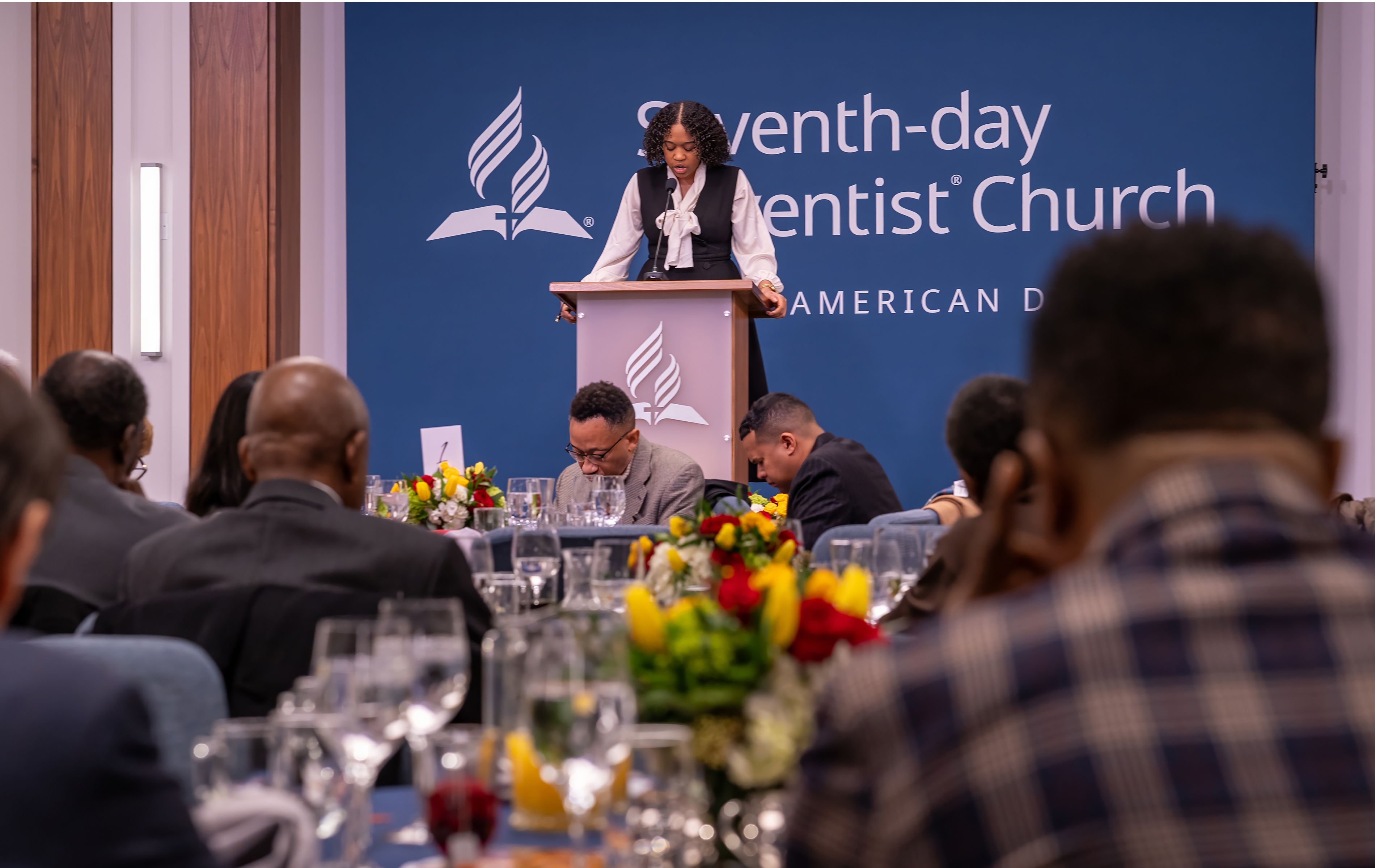 Black woman praying in front of a crowd in conference room. 