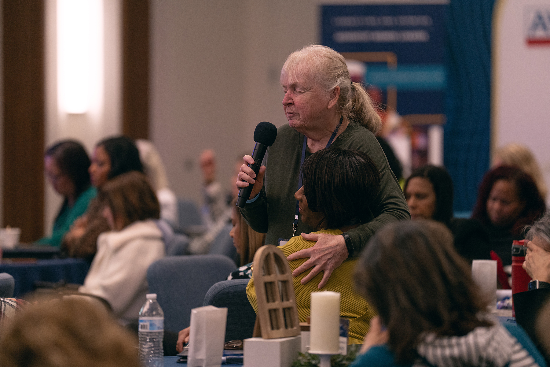 A white woman standing and speaking into a mic hugs a black woman who is seated. Both have their eyes closed