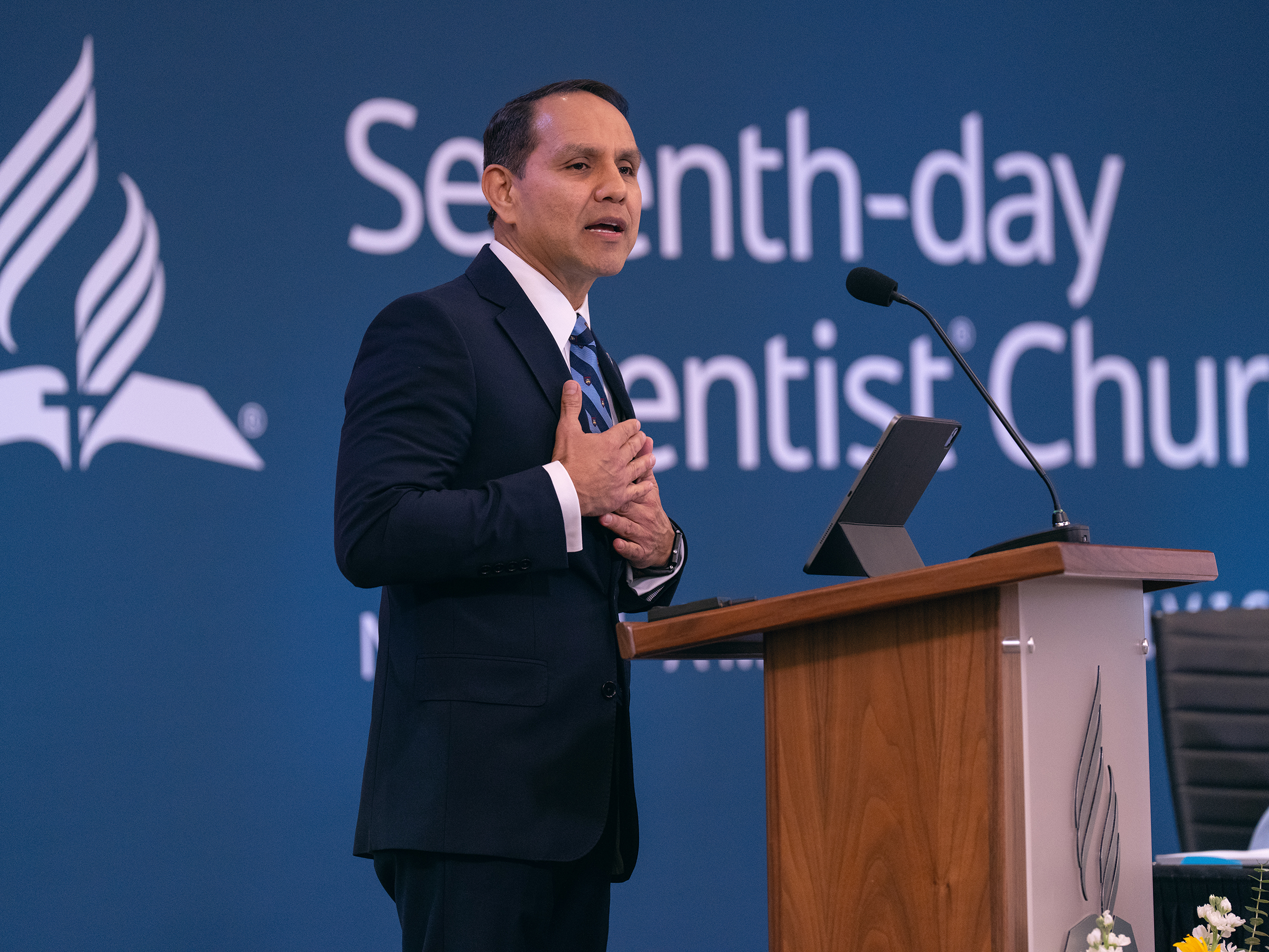 A Hispanic man does a presentation in a suit, hands on chest with an earnest expression.