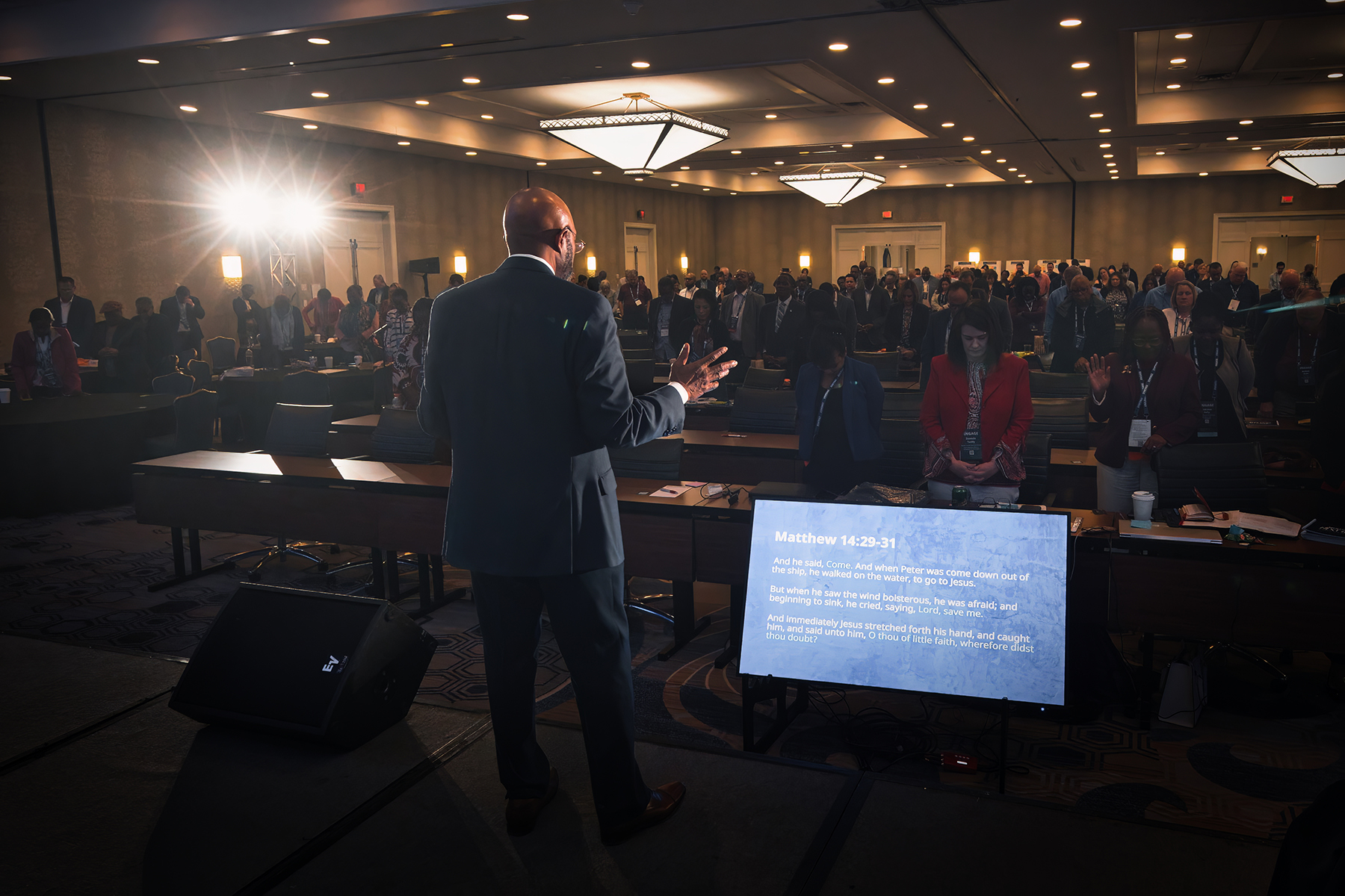  A man stands in front of a big crowd speaking. He is visible with lights and the crowd is dark