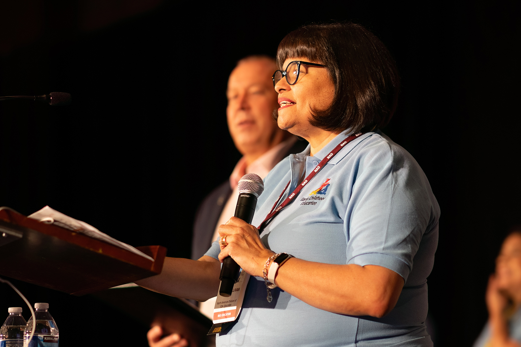 A white woman is in the foreground, speaking, while a white man is blurred in the background.