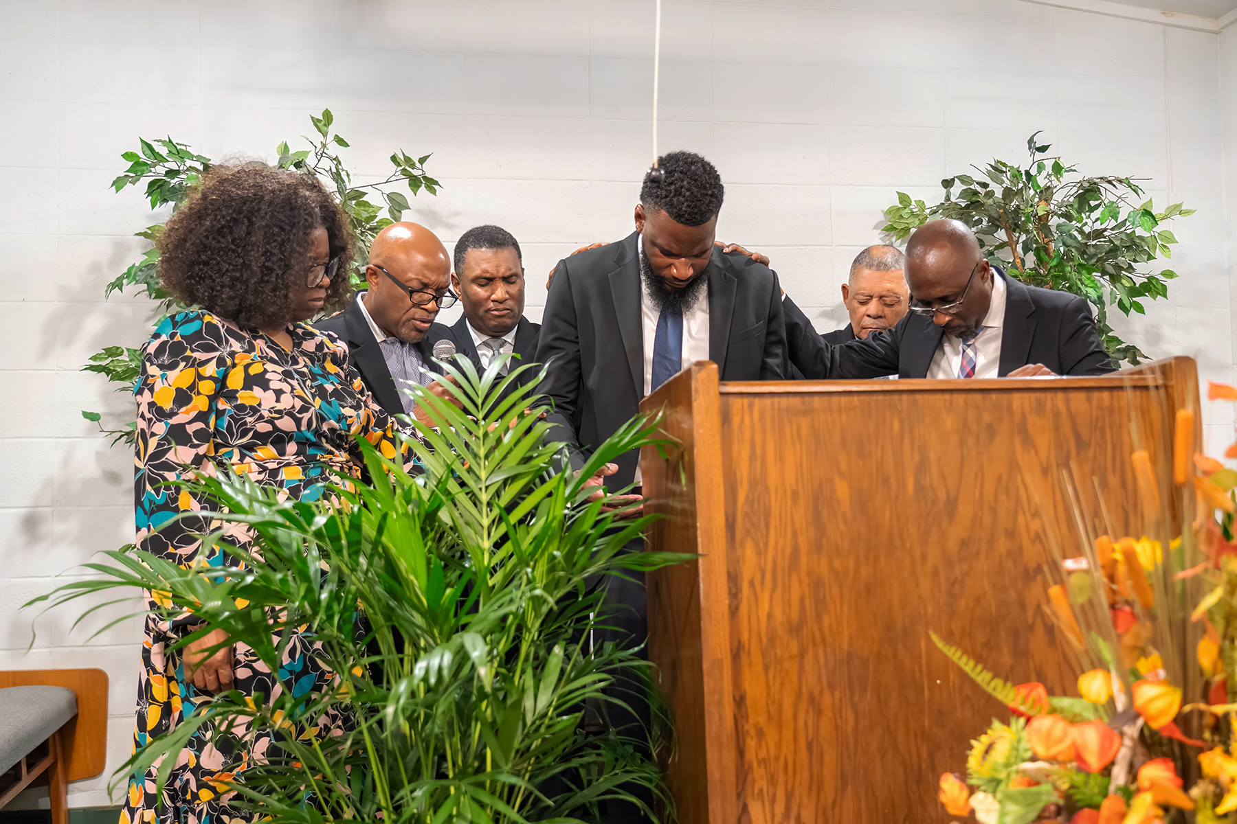 Photo of several men and a woman on a church pulpit, praying over the pastor in the middle.