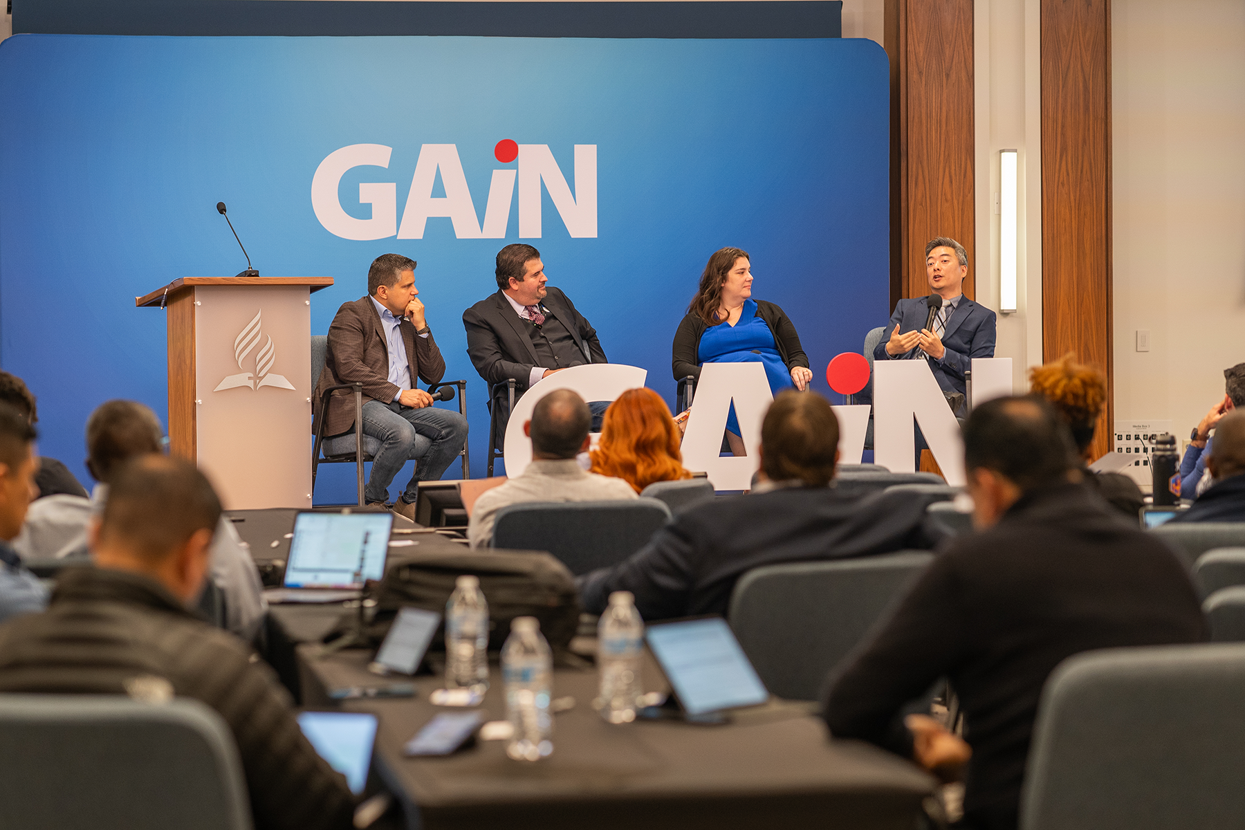 Left to right: two white men, white woman, and Asian man, seated, engage in a panel discussion at a conference.