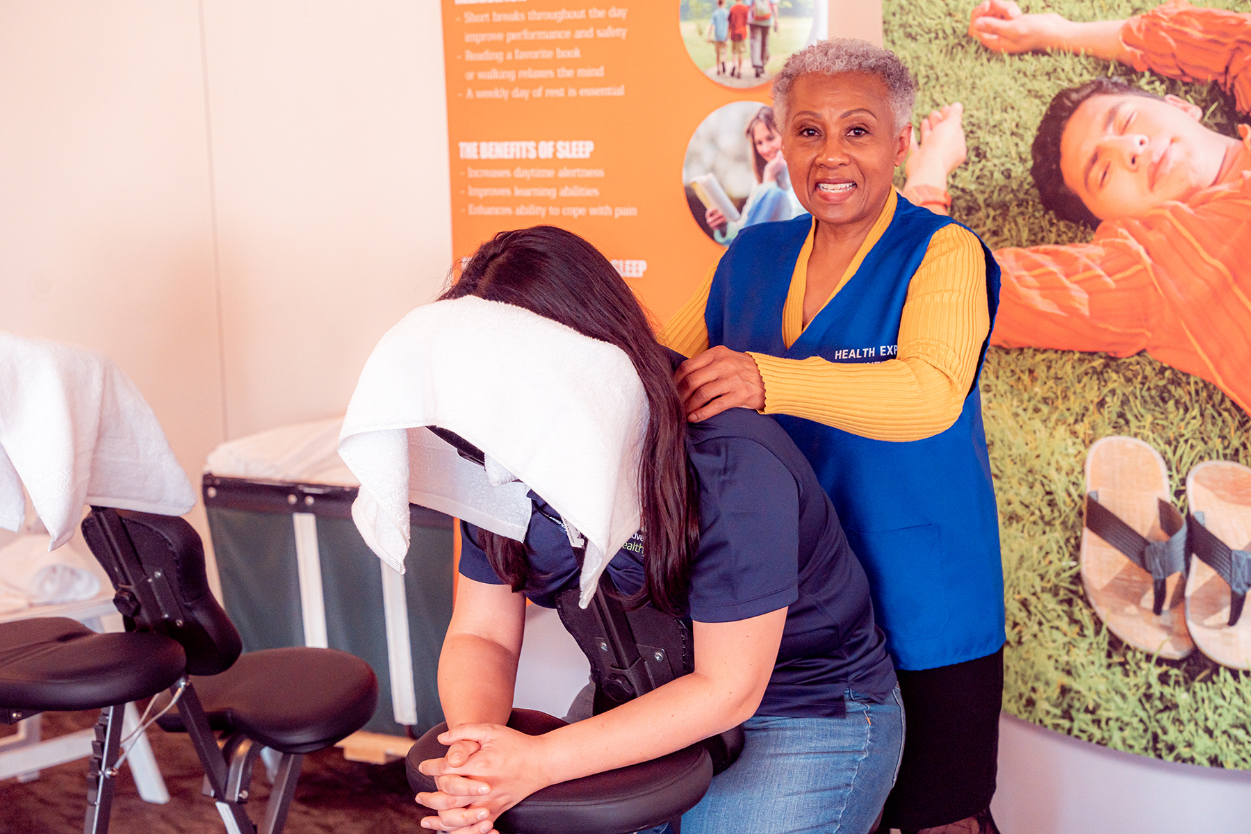 A woman, standing, massages the shoulders of another woman, seated. The client's face is covered with a towel.