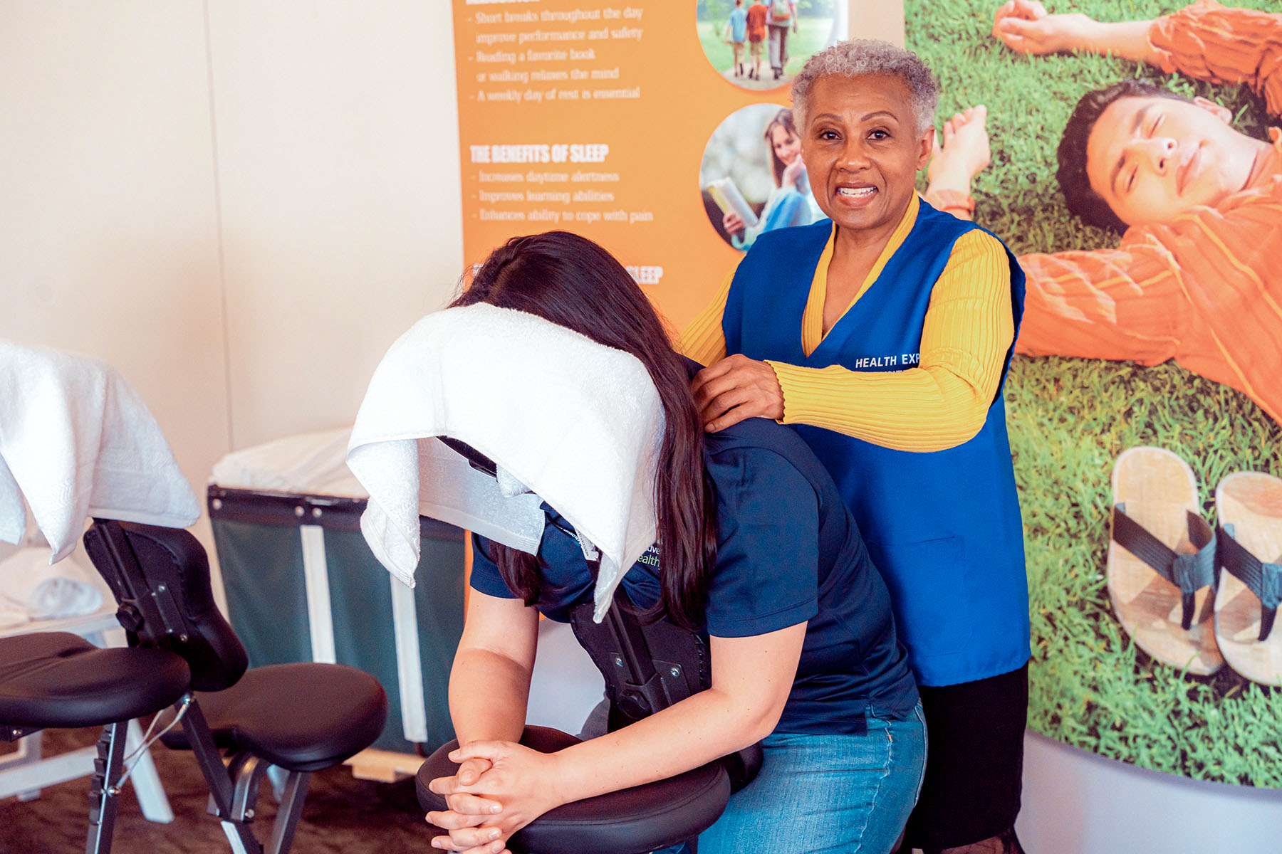 A woman, standing, massages the shoulders of another woman, seated. The client's face is covered with a towel. 