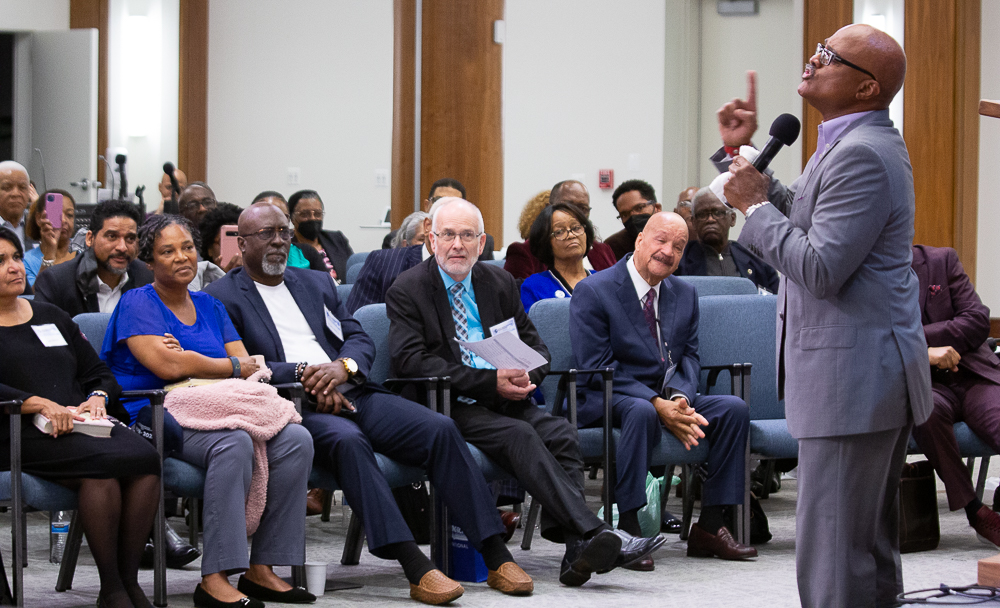 A Black man preaches in front of a diverse crowd of men and women