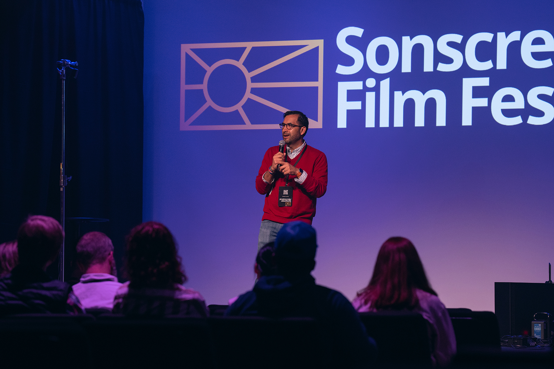 A man stands on a stage speaking in front of a screen with a backdrop reading "Sonscreen Film Festival."