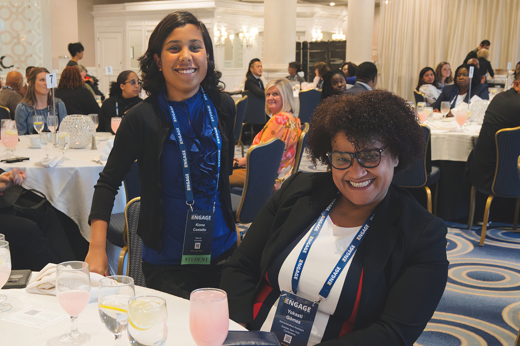 Two women, one standing beside a round table and one seated, smile as they engage at a conference