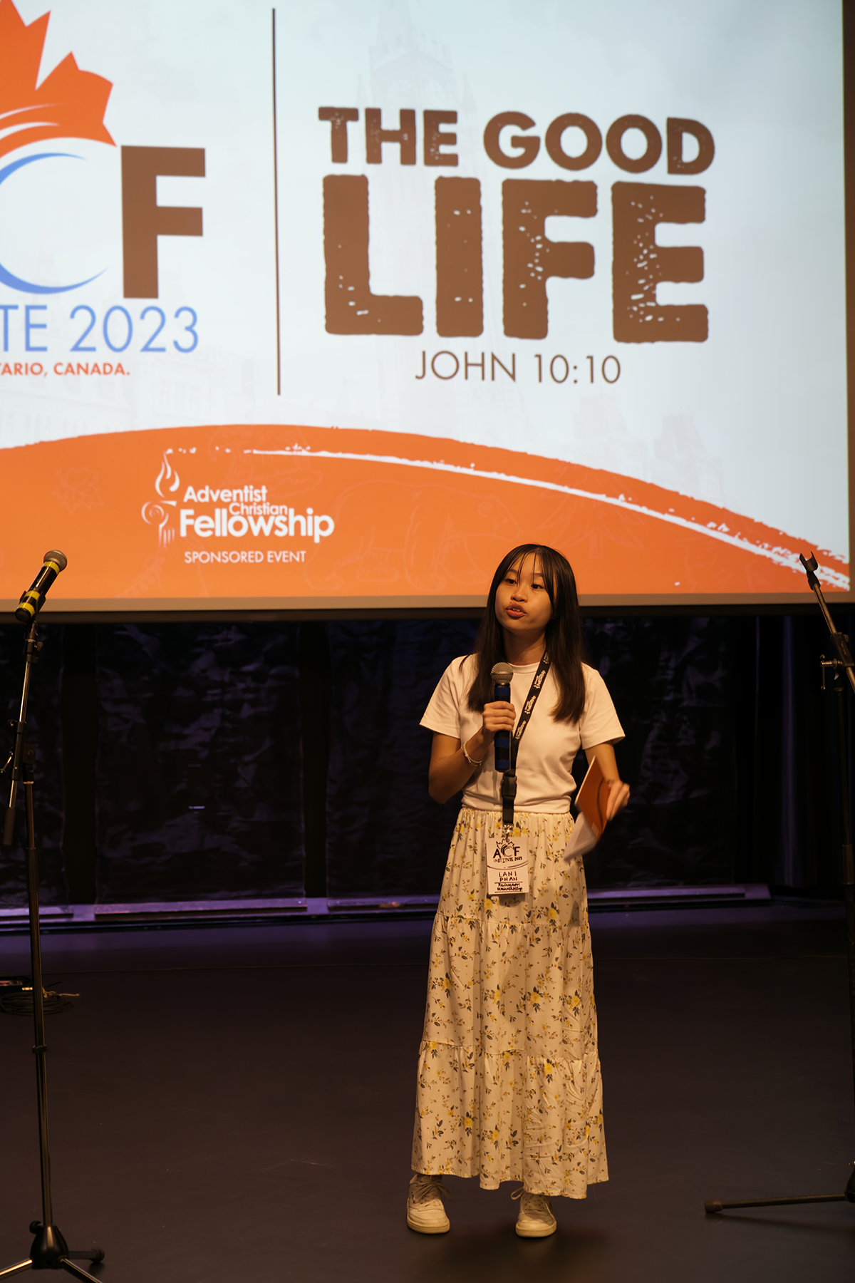 Asian young woman holding a notebook stands on a darkened stage speaking