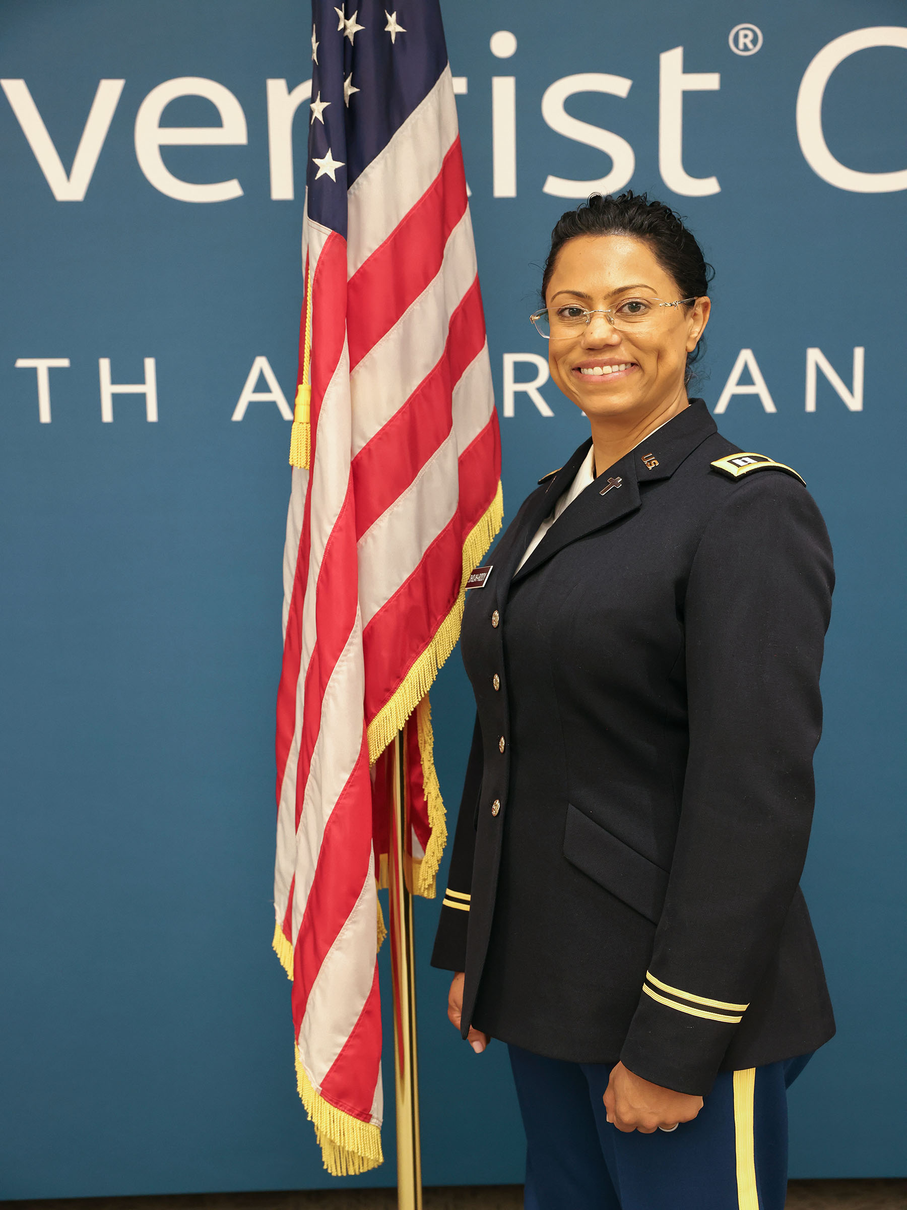 Woman in military uniform smiling as she stands next to the American flag.