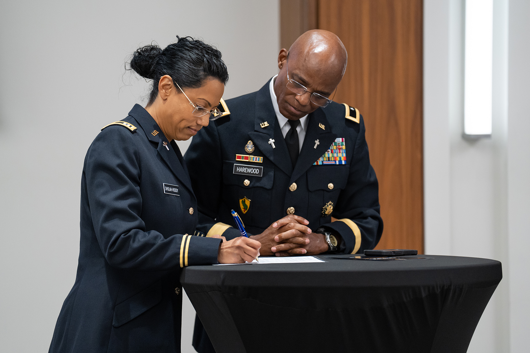 A lady with tanned skin in military uniform signs a paper as a black man, also in uniform, watches.