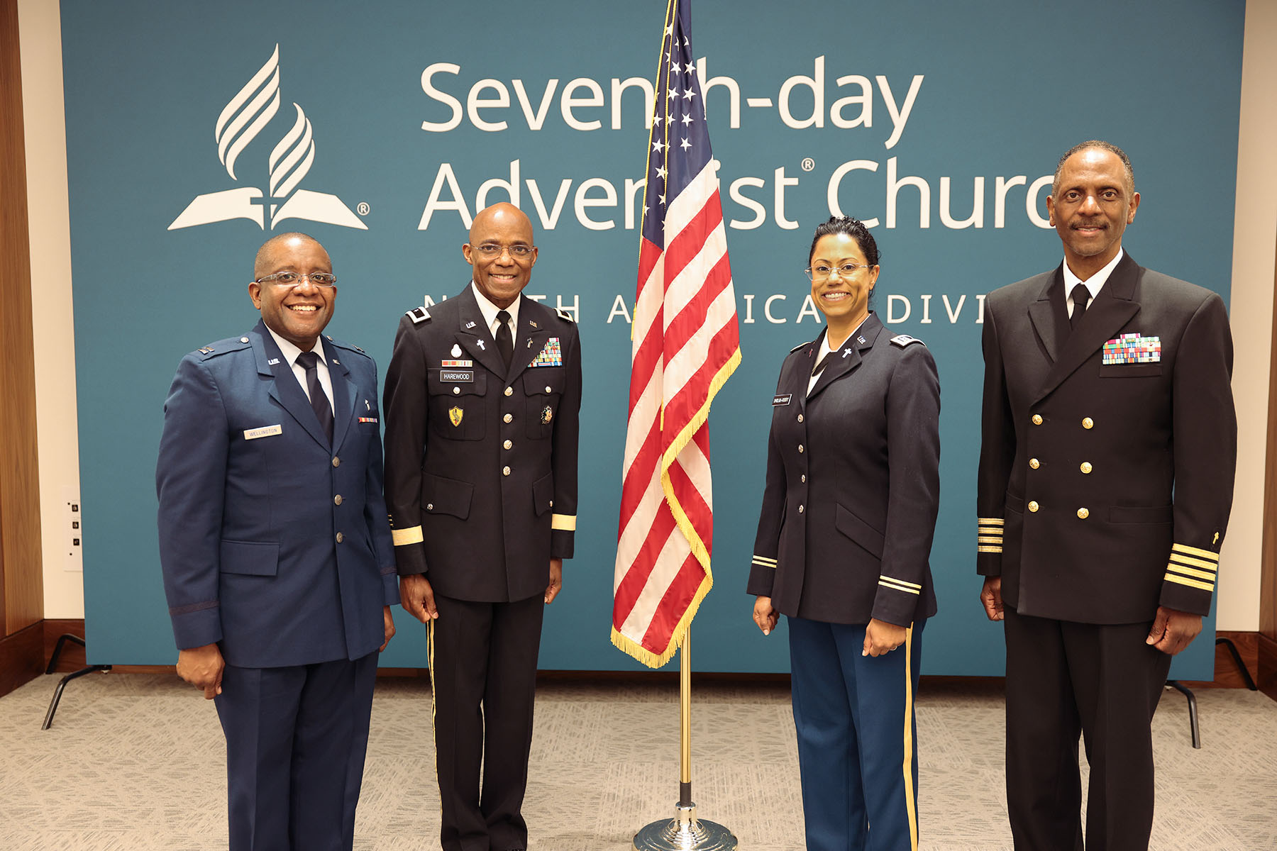 Four individuals in uniform, one woman with tanned skin and three black men, stand proudly with the U.S. flag in between them. 