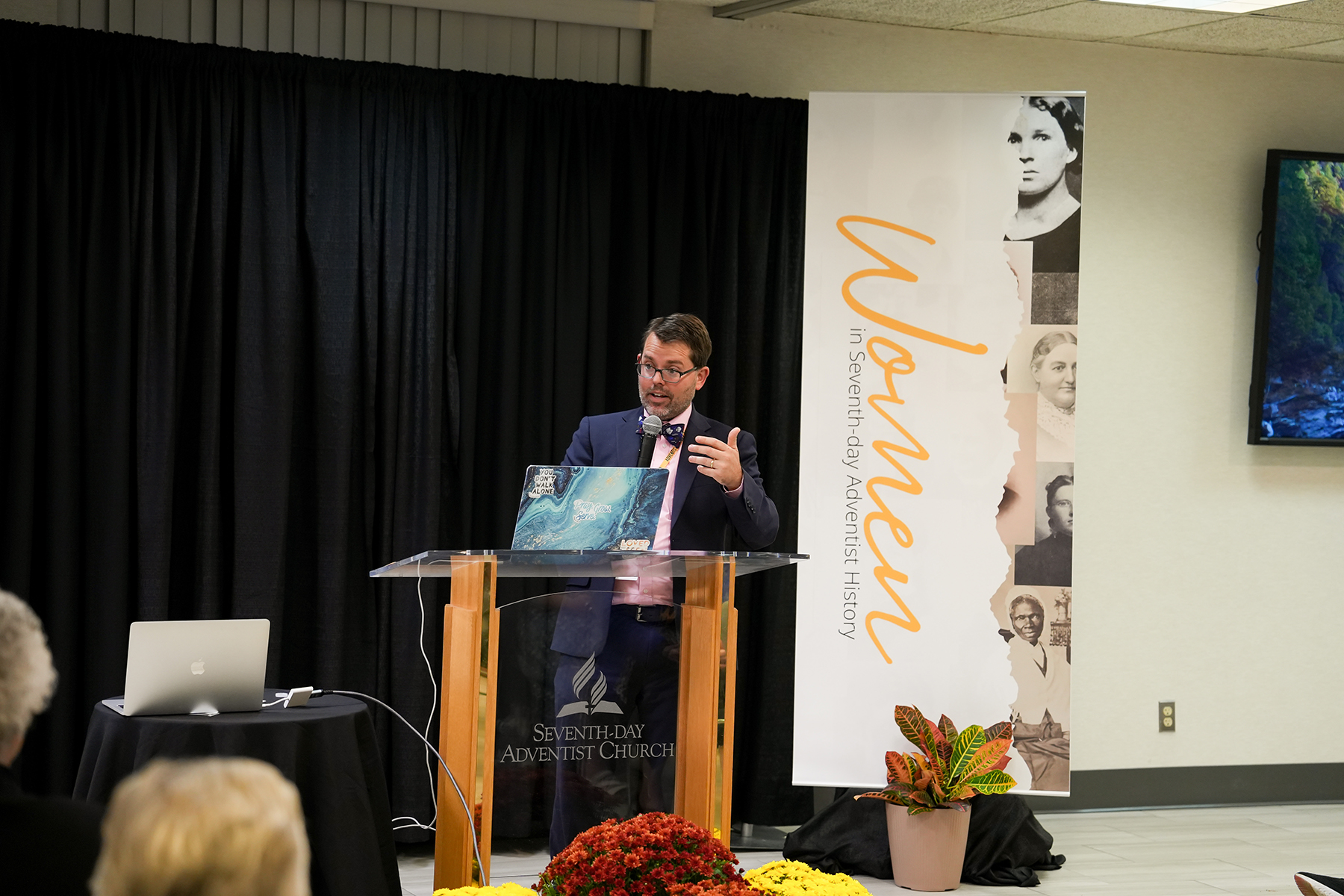 Caucasian giving a speech to a crowd from behind a podium. There is a sign reading "Women in Adventist History Conference" beside the podium.